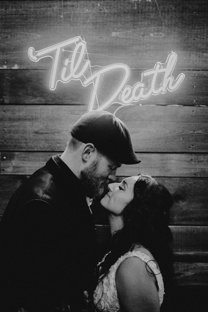 black and white photo halloween wedding bride and groom kissing with neon Til Death sign in the background