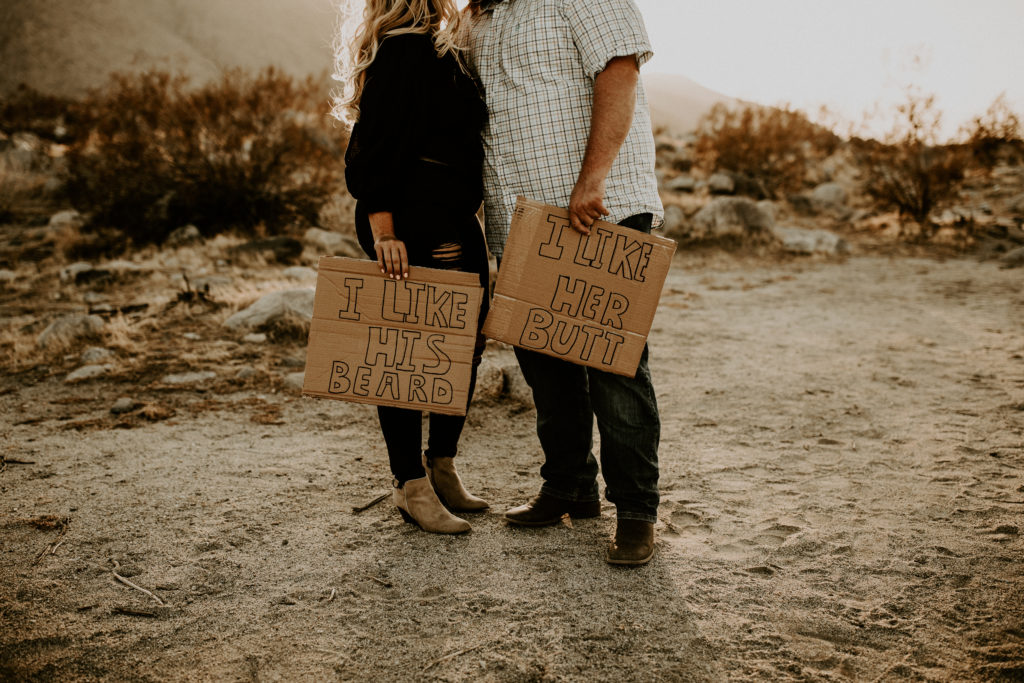 couple holding card board signs while taking desert engagement photos in Palm Springs