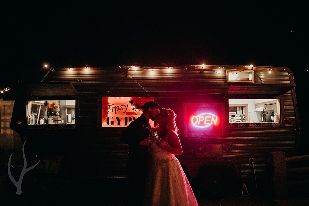 bride and groom kiss in front of back lit bar