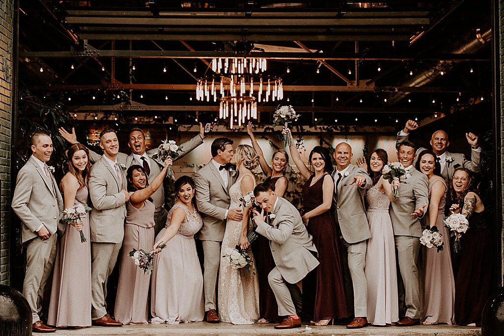 large bridal party in tan tuxes and blush bridesmaid dresses at entrance of rustic wedding venue