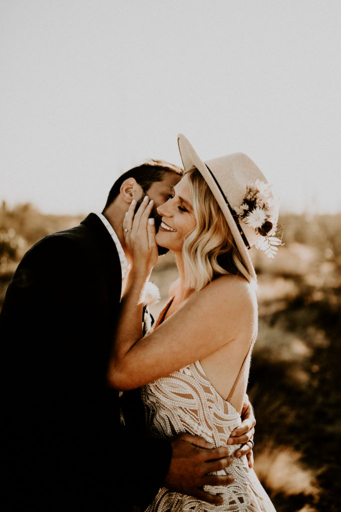 groom kissing bride with hat on with desert in background