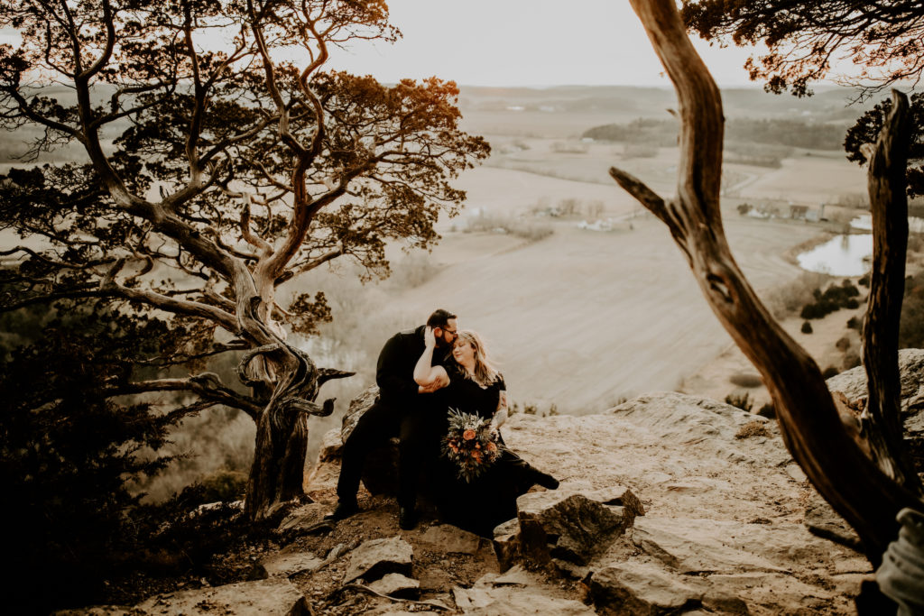 couple in all black sitting on large rock near a unique tree overlooking the valley