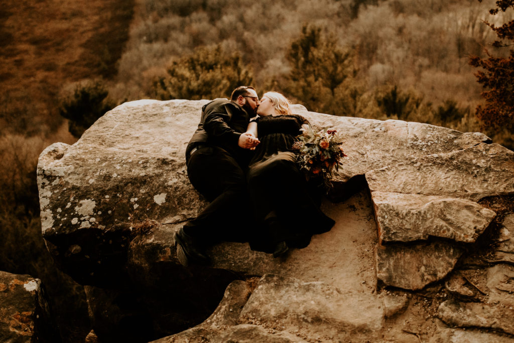 newlywed couple in all black laying on large rock above a open valley