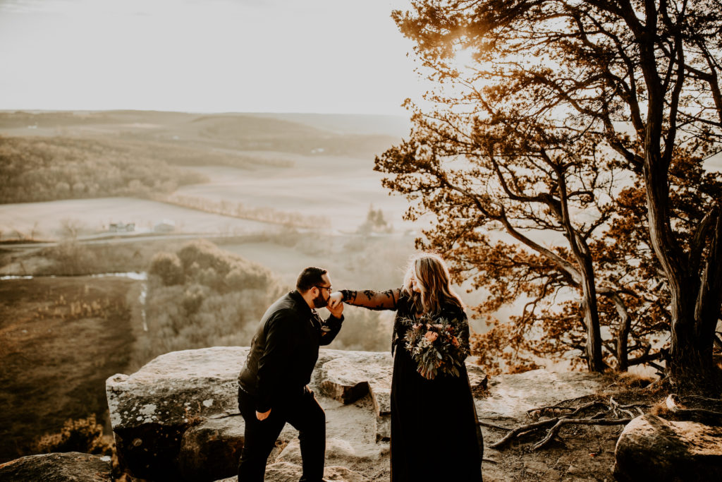 husband kissing wife's hand while standing on a large rock overlooking the valley