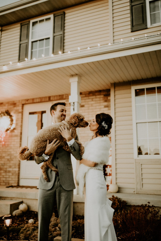 bride and groom kissing dog in front of house at their backyard wedding