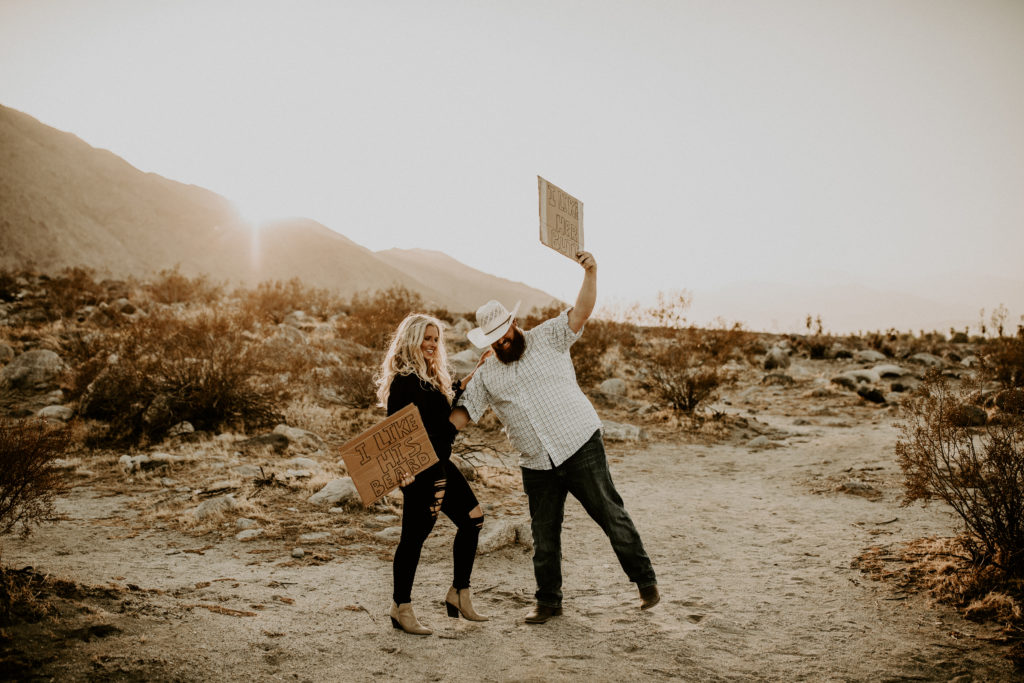 couple holding cardboard signs in the middle of the desert with mountains in the background